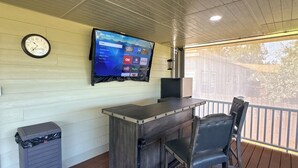 A covered outdoor seating area with a TV mounted on the wall displaying various streaming services, a clock, and a trash can. - Enjoyable 3-bedroom house with Jacuzzi, Mini Golf and Cantal AC in Canyon Lake. (Canyon Lake)