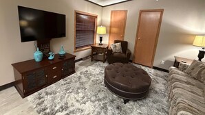 Un salon comprenant un téléviseur à écran plat, un pouf marron, un canapé, un meuble en bois, une lampe et une fenêtre avec des stores.