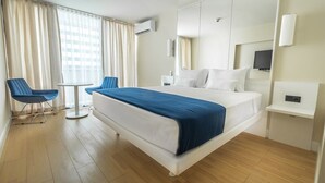 A hotel room with a large bed, a small round table, two blue chairs, a TV, and a wall-mounted air conditioning unit. - Orbi City Panorama Sea View Studios (Batumi)
