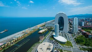 A modern hotel with a distinctive arch-shaped entrance, located near a waterfront with a marina and a road leading to the city center. - Orbi City Panorama Sea View Studios (Batumi)