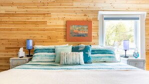 A bedroom with a wooden wall, a bed with striped bedding, a nightstand with a lamp, and a window with a view of trees. - Hammy on a Rye 2 Hammondsport NY (Hammondsport)