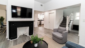 A modern living room with a flat-screen TV, a fireplace, a grey armchair, and a staircase leading to a kitchen area.