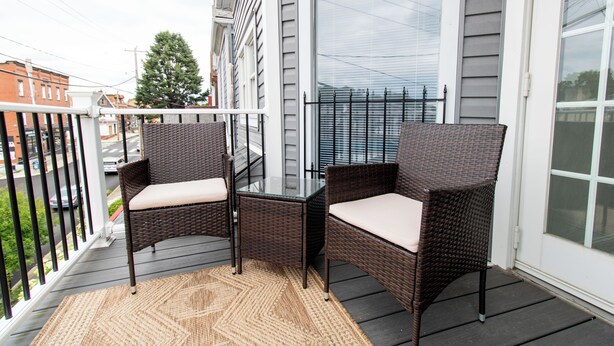A balcony with wicker furniture and a glass table, overlooking a street with buildings and a tree.