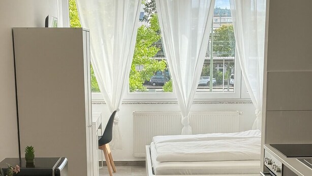 Une cuisine moderne avec un lit, un coin repas et une vue sur la rue à travers des rideaux transparents.