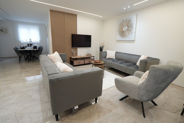 A modern living room with a grey sofa, a grey armchair, a flat-screen TV, and a dining area with a table and chairs.
