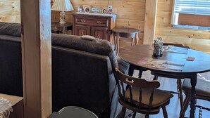 Interior acolhedor de cabana com teto de madeira, uma TV de tela plana na parede, uma mesa de jantar com cadeiras, um sofá e uma lixeira.