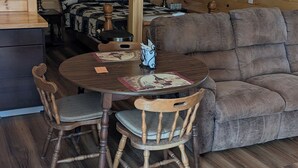 Interior acolhedor de uma cabana com mesa de jantar de madeira, duas cadeiras, um sofá e um ventilador de teto.