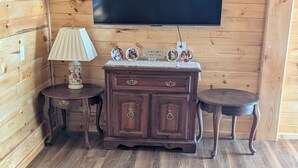 Um quarto com painéis de madeira, uma televisão de tela plana na parede, um armário de madeira escura com gavetas, duas mesinhas de cabeceira e um sofá grande.