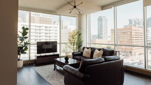 Una sala de estar moderna con un sofá de cuero, una mesa de centro y un televisor de pantalla plana. Ventanas amplias ofrecen vista a la ciudad.