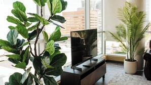 Una sala moderna con un gran ventanal, un televisor de pantalla plana sobre un mueble de madera oscura y dos plantas en macetas.