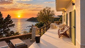 A terrace with a view of the sea at sunset, outdoor furniture, and a building with shutters.