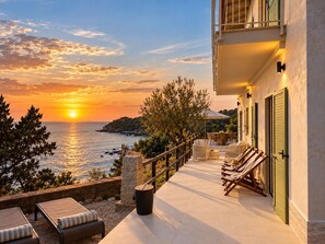 A terrace with a view of the sea at sunset, outdoor furniture, and a building with shutters.