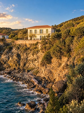 A coastal building with a red-tiled roof, situated on a rocky hillside with a view of the sea. - Roc Villa - 2 Private Beach Accesses  (Rosignano Marittimo)