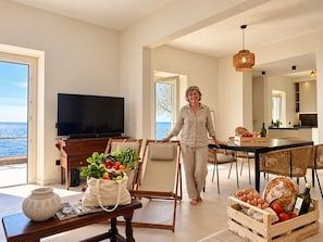 A spacious living area with a television, dining table, and chairs. A woman is walking through the room. - Roc Villa - 2 Private Beach Accesses  (Rosignano Marittimo)