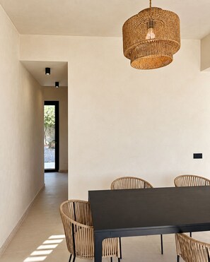 A modern dining area with a dark table and woven chairs, a woven pendant light, and a view through an open doorway. - Roc Villa - 2 Private Beach Accesses  (Rosignano Marittimo)