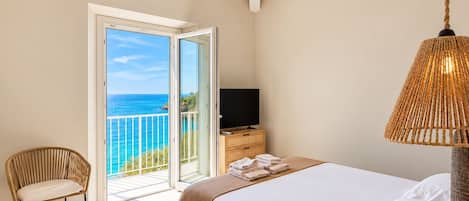A bedroom with a bed, a television, a chair, and a view of the sea through a glass door.