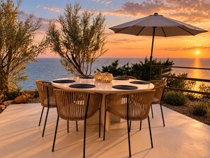 A round outdoor dining table with six chairs, a large umbrella, and a view of the sea at sunset. - Roc Villa - 2 Private Beach Accesses  (Rosignano Marittimo)