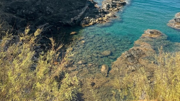 A coastal landscape with clear blue waters, rocky terrain, and sparse vegetation.