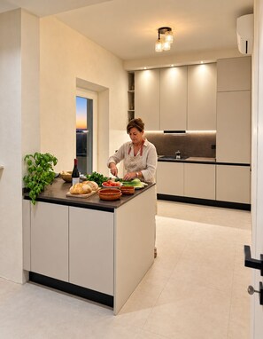 A modern kitchen with a central island, a person preparing food, and a view of the sunset through a window. - Roc Villa - 2 Private Beach Accesses (Rosignano Marittimo)