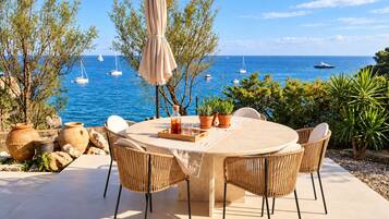 A round table with four chairs, a tablecloth, and a bottle of water, set on a patio overlooking the sea with boats.