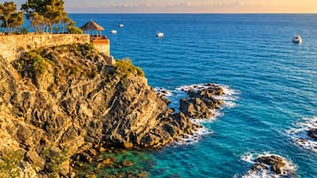 A coastal landscape with rocky cliffs, clear blue sea, and a small structure on the cliff.