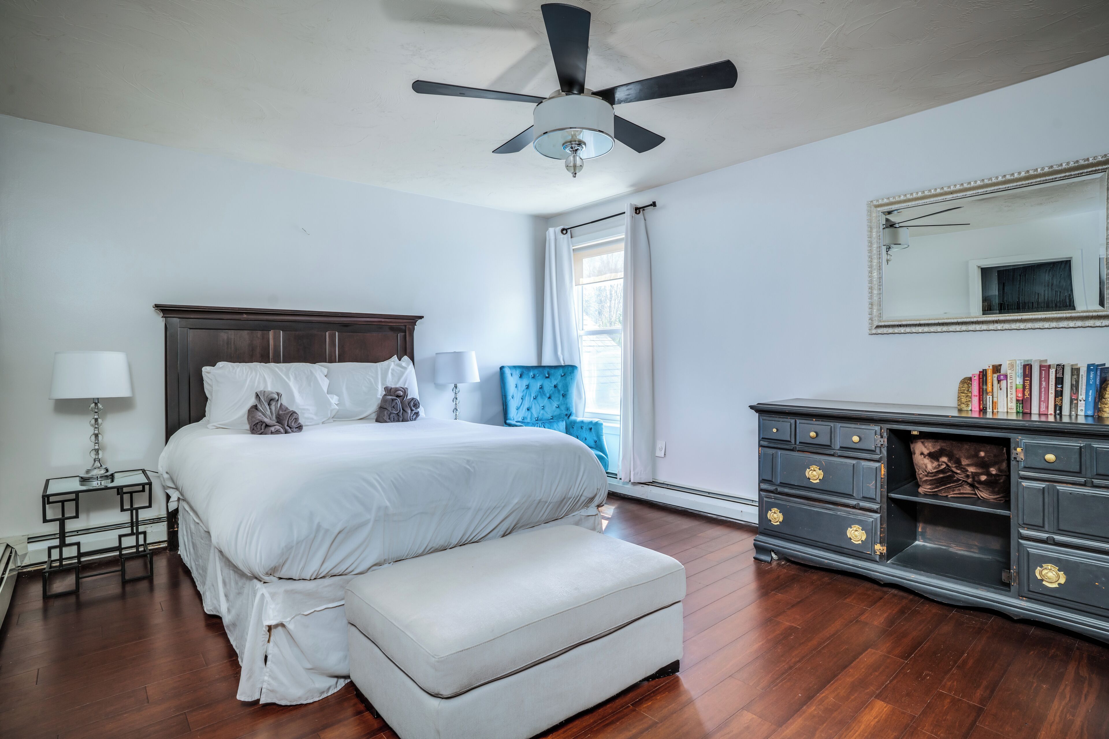 A bedroom with a large bed, a bedside table with a lamp, a dresser with drawers, a ceiling fan, and a window with curtains.