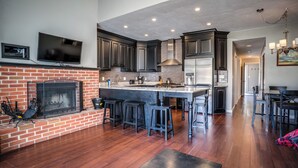 A modern kitchen with dark cabinetry, a central island with bar stools, a fireplace, and a television mounted above it. - Tenney Mountain Retreat, Plymouth & Rumney, NH (Plymouth)