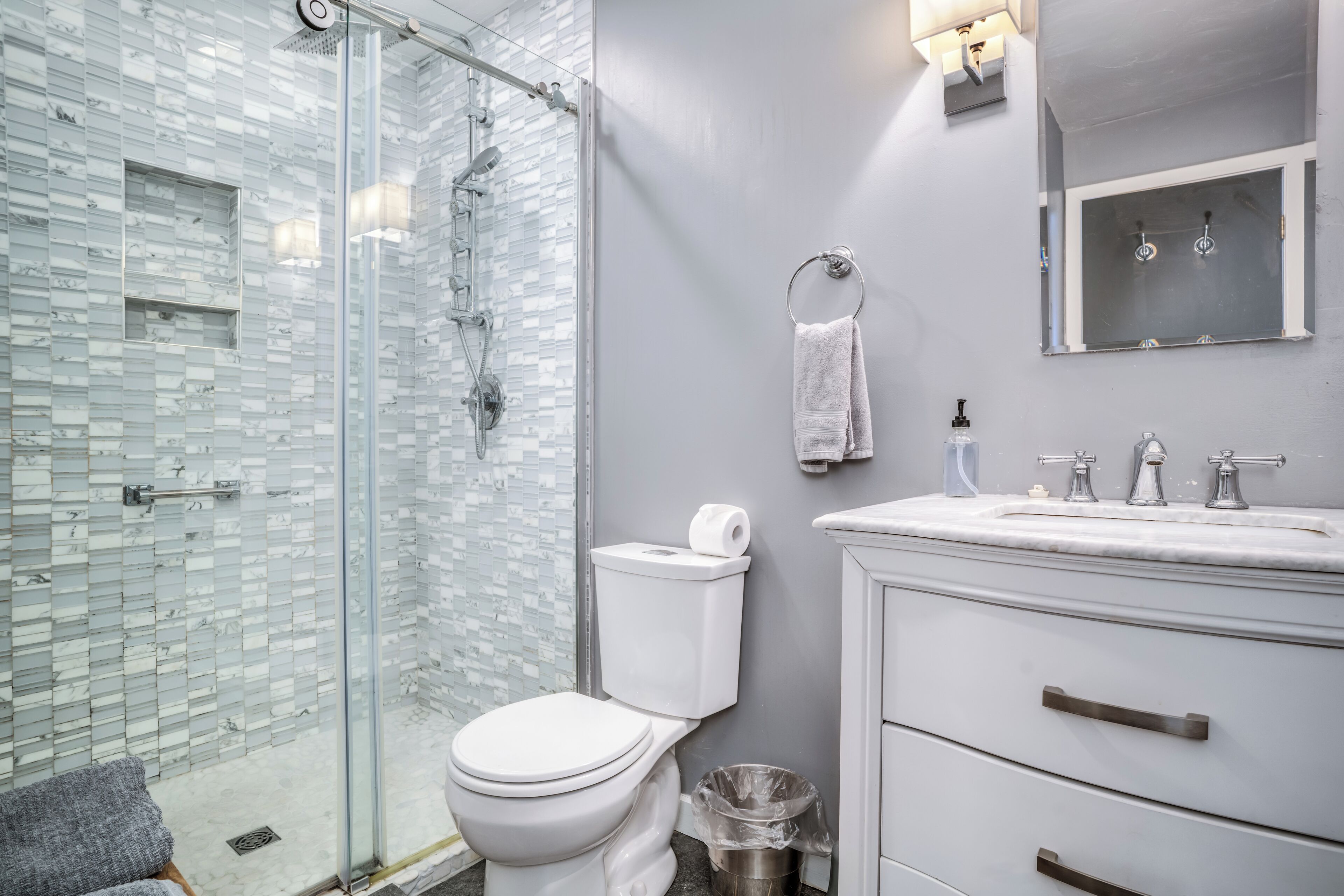 A modern bathroom with a glass shower enclosure, a white toilet, a sink with a mirror, and a trash can.