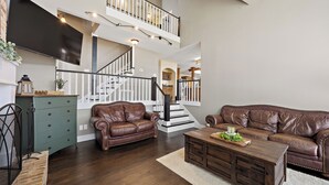 A spacious living room with a staircase, leather sofas, a wooden coffee table, and a flat-screen TV mounted on the wall.