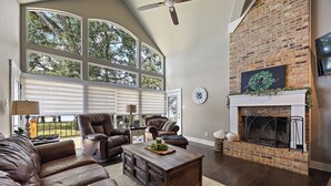 A spacious living room with a large window, leather sofas, a fireplace, and a brick accent wall.
