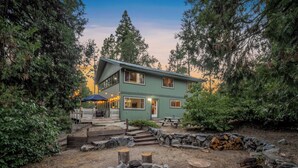 A two-story house with a patio area, surrounded by trees and a fire pit.