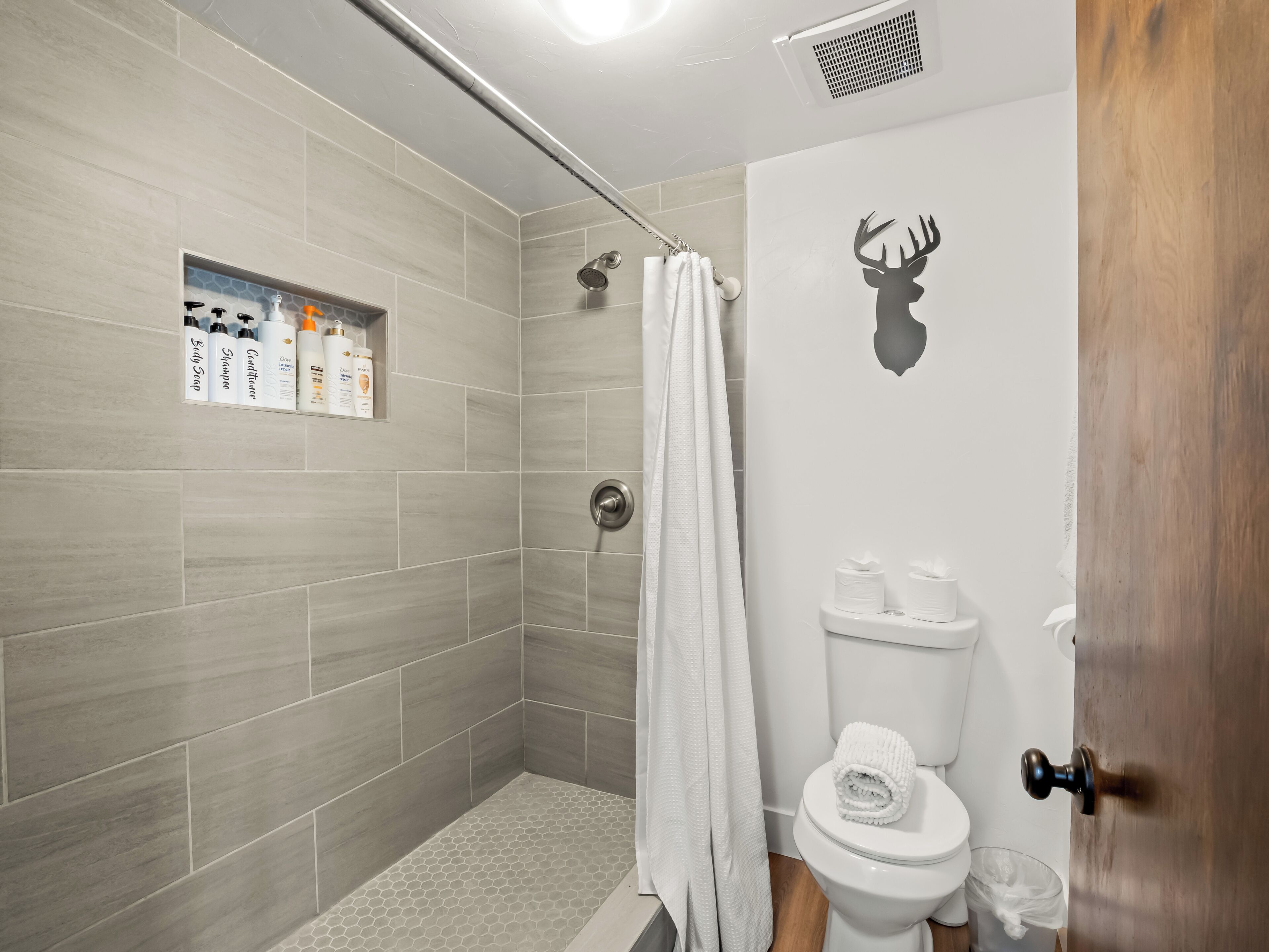 A bathroom with grey tiles, a shower, a toilet, and a shelf with toiletries.