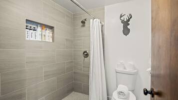 A bathroom with grey tiles, a shower, a toilet, and a shelf with toiletries.