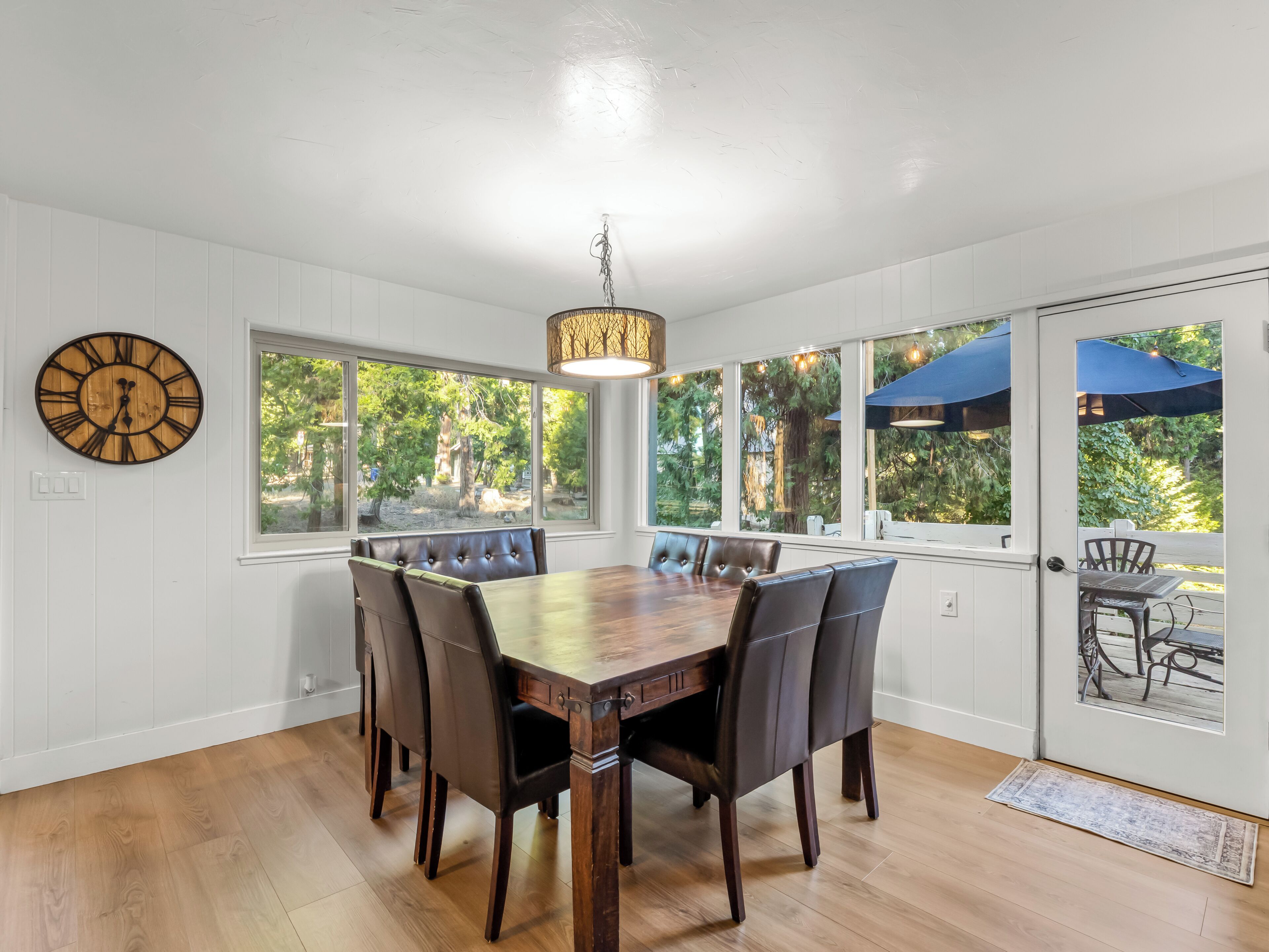 A dining area with a wooden table and chairs, large windows, and a wall clock.