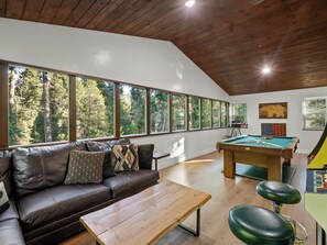 Une pièce avec un plafond en bois, une table de billard, un canapé en cuir et de grandes fenêtres donnant sur une forêt.