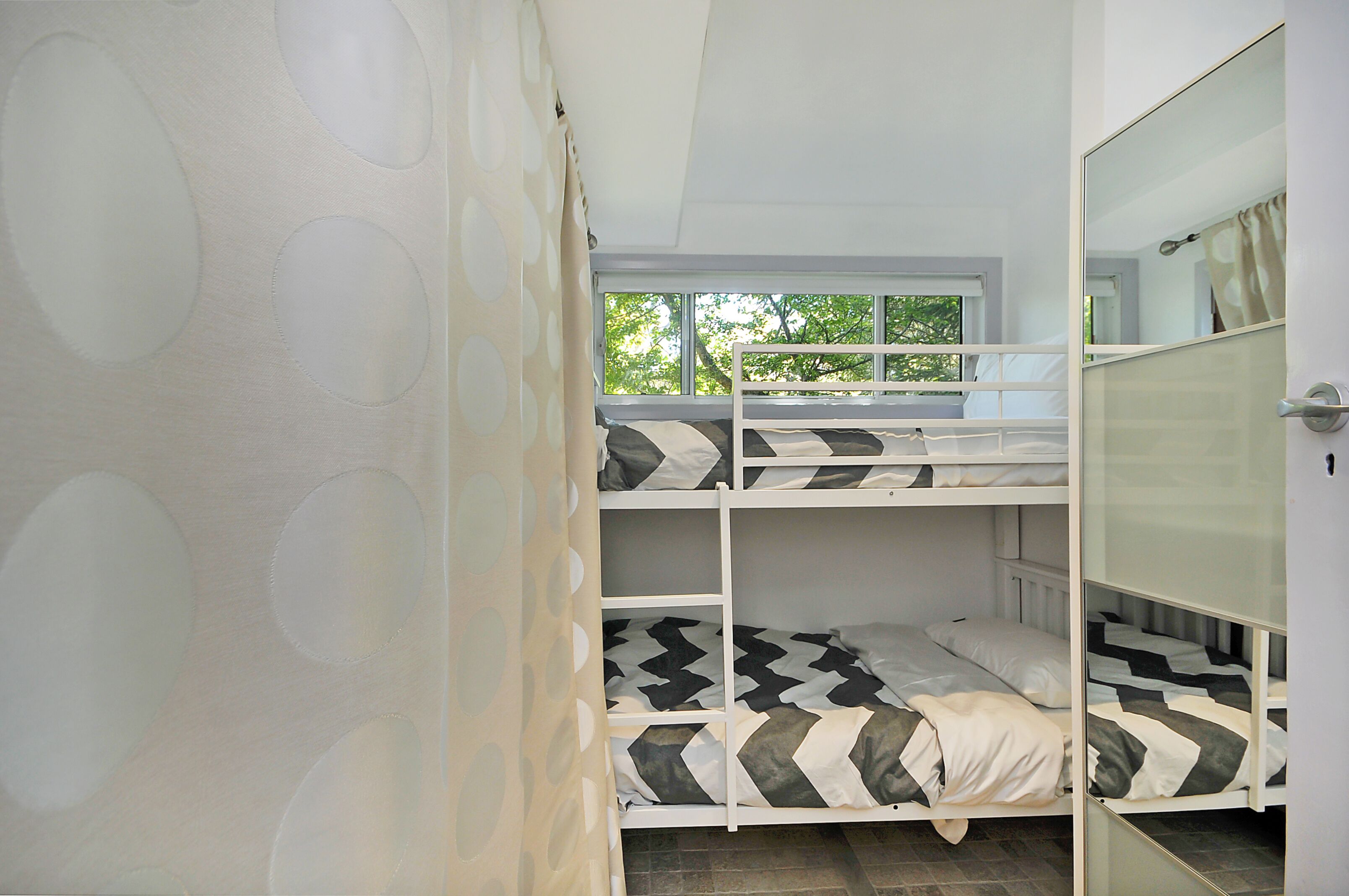 Une chambre avec un lit superposé, une fenêtre, des rideaux blancs et un miroir.