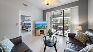 A modern living room with a glass coffee table, leather sofas, a flat-screen TV, and a balcony with outdoor seating.