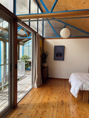 A bedroom with wooden flooring, a bed, a potted plant, a framed picture, and a view of the outdoors through a sliding glass door. - 'The Dogbox' Architectural home in Central Whanganui. (Whanganui)