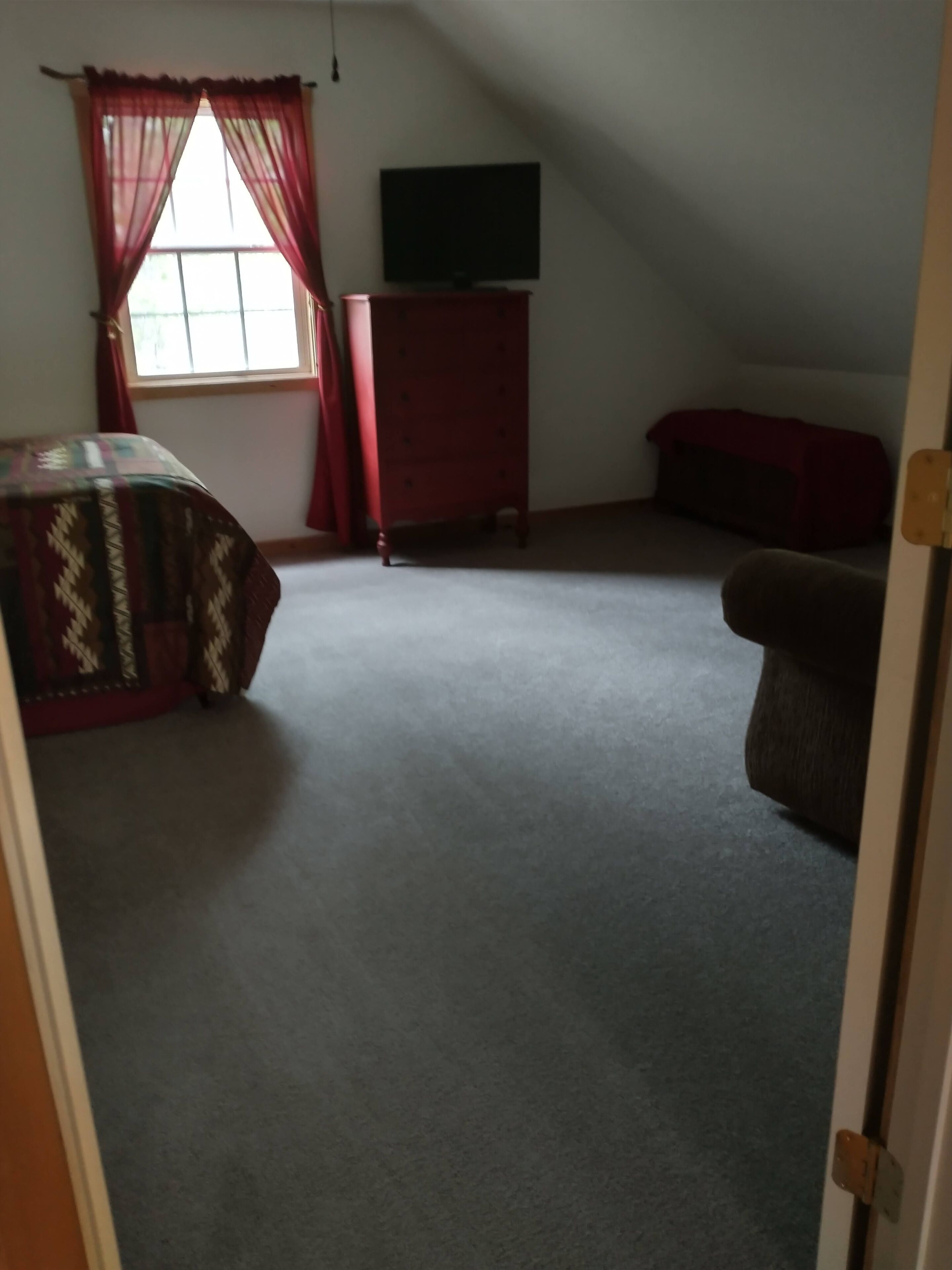 A room with a sloped ceiling, a television mounted on the wall, a red dresser, a patterned bedspread, and a red couch.