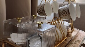 Een transparante glazen salontafel met een gouden voet en een dienblad met een set witte kopjes met gouden handgrepen.