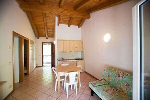 A room with a wooden ceiling, a dining table with chairs, a kitchenette, and a sofa.