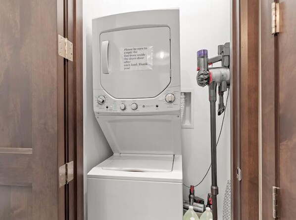 Un appareil compact, blanc, comprenant une machine à laver et un sèche-linge, conçu pour être fixé au mur, avec un panneau de commande et un tuyau.