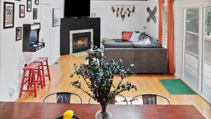 A modern living room with a fireplace, a dining table with a vase of flowers, and a large window. - Modern Chalet with Hot Tub, Fire Pit & Mountain Views in Basye, VA (Basye)