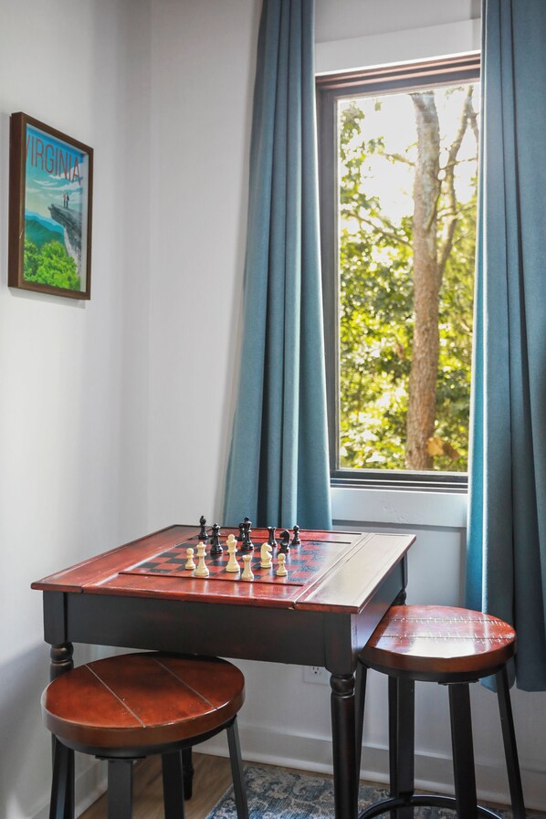 Ein Schachtisch mit zwei Hockern, ein Fenster mit Vorhängen und ein Gemälde an der Wand.