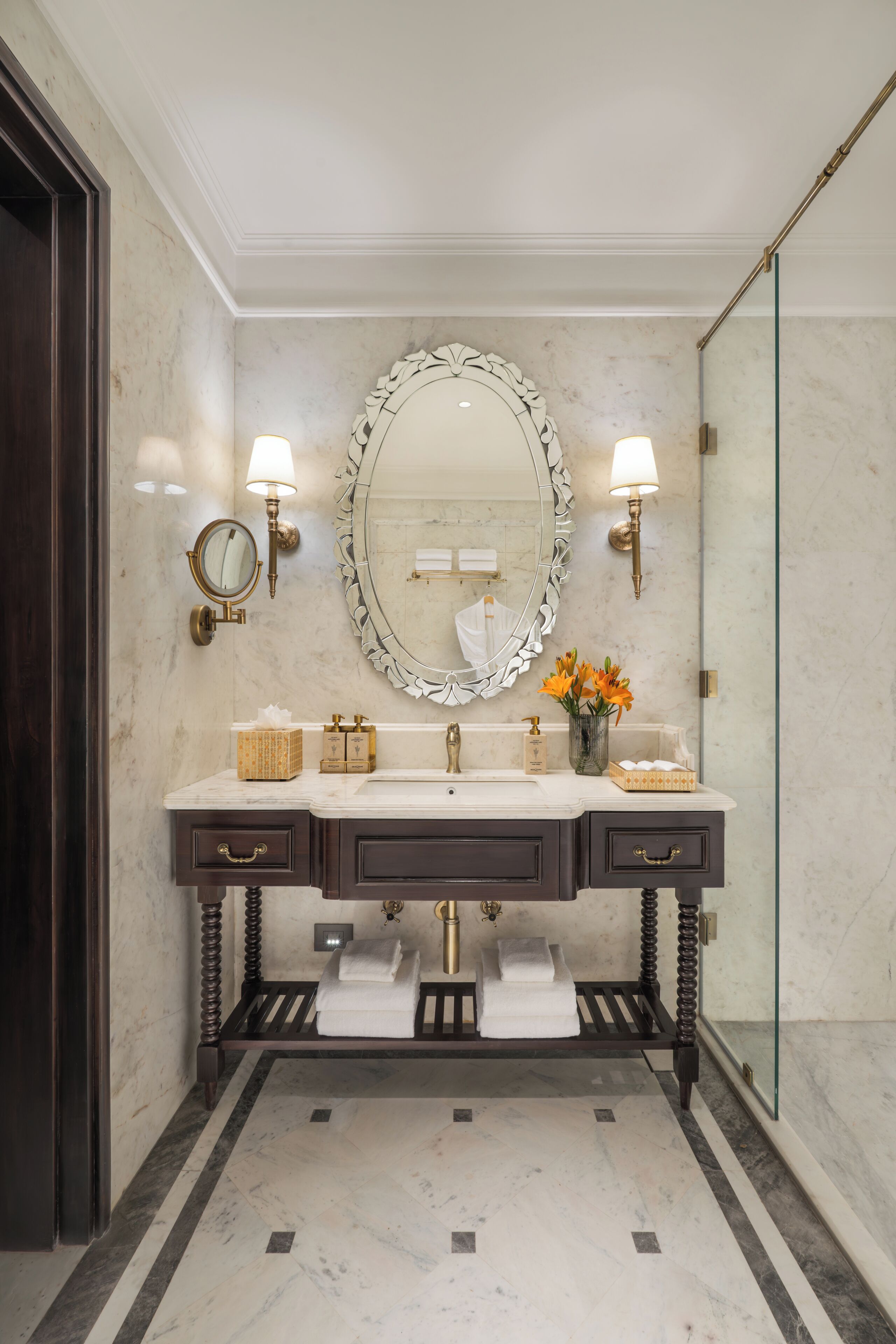 A bathroom with a large mirror, two wall-mounted lights, a sink with a towel rack, and a glass shower enclosure.
