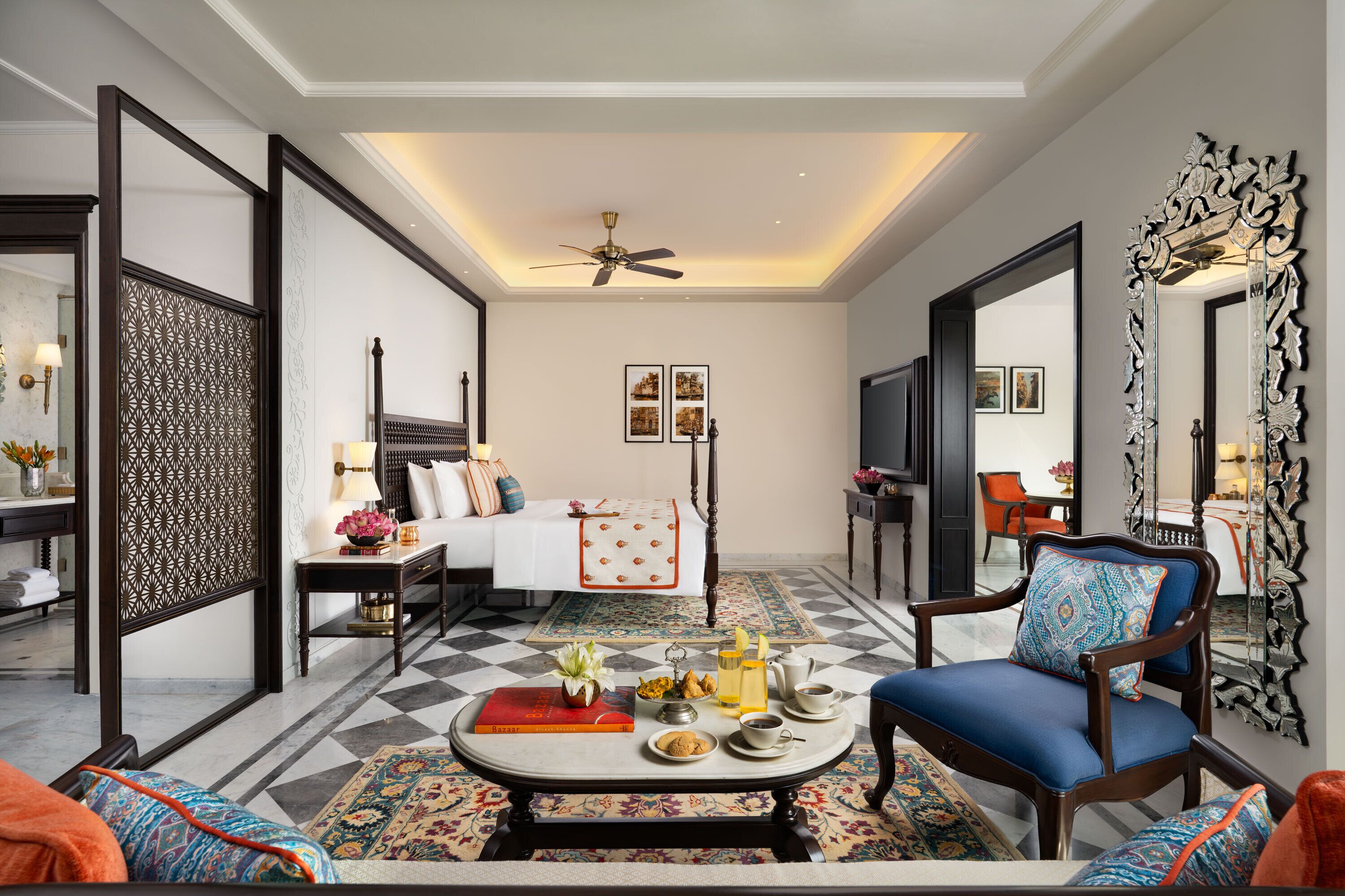 A hotel room with a checkered floor, a bed with a white and orange bedspread, a sitting area with a coffee table, and a large mirror with an ornate frame.