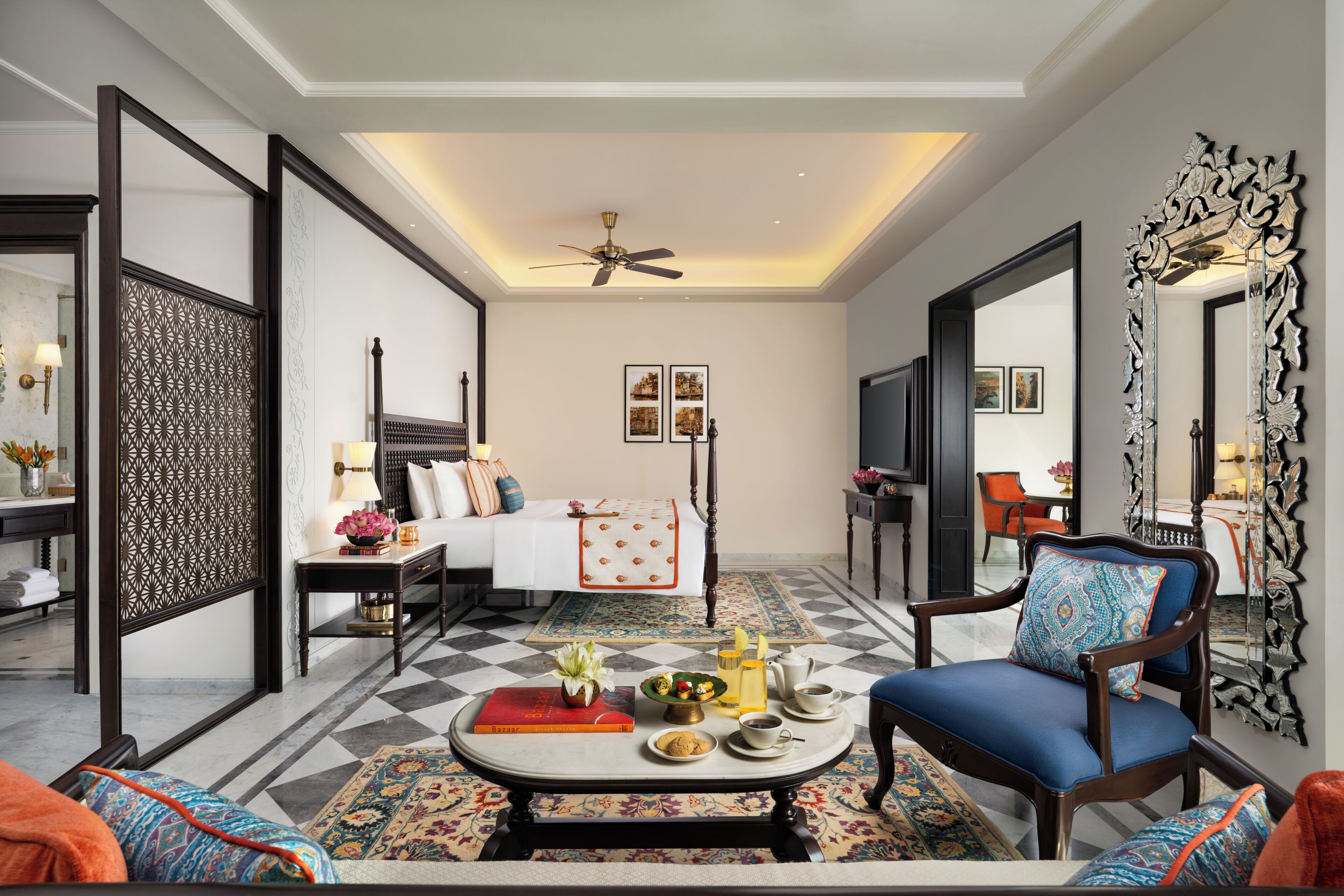 A hotel room with a bed, a sitting area with a coffee table, a chair, a large mirror, and a ceiling fan.