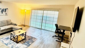 Een woonkamer met een bank, een salontafel, een aan de muur bevestigde televisie en een raam met zonwering.