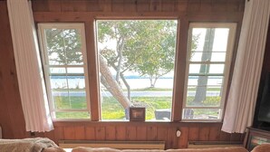A room with a wooden paneled wall, a large window with curtains, and a brown sofa. - Charming Lakefront Cottage (Bear Lake)