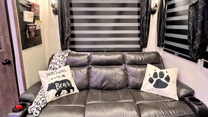 A room with a black leather sofa, two pillows with animal prints, a wall-mounted shelf with books, and a window with black curtains. - The Bear Retreat at Venus (Highlands County)